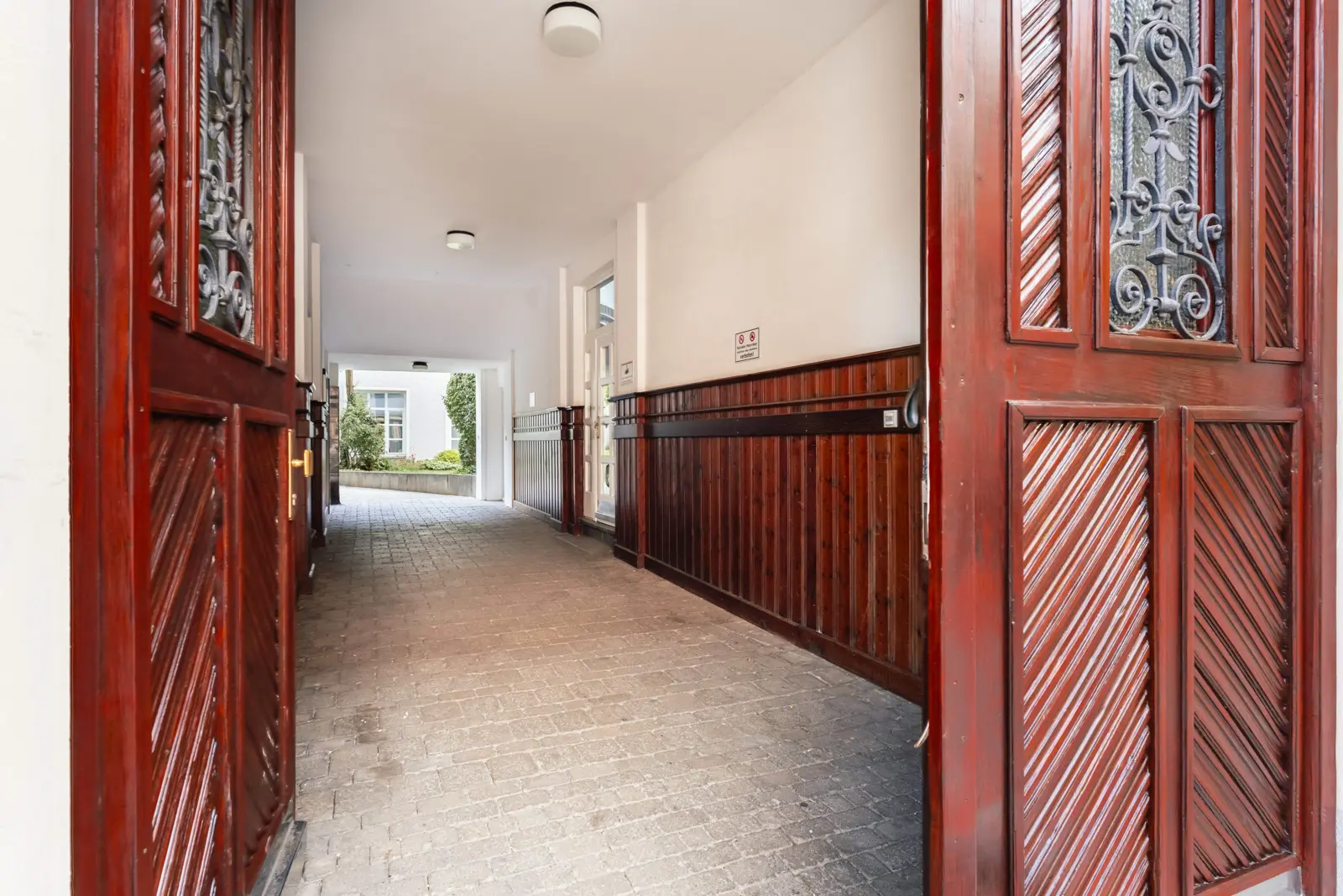 Open wooden doors lead to a covered passageway with white walls and dark wood paneling, ending at a courtyard with greenery visible in the distance.