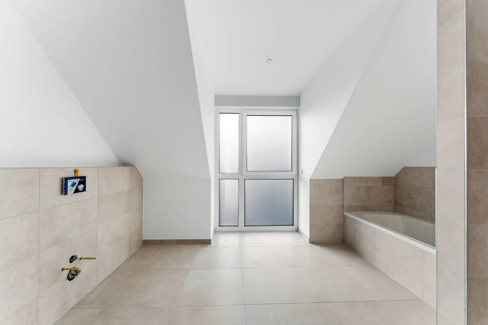 Modern bathroom under a sloped ceiling with beige tiles, frosted glass windows, and a built-in bathtub on the right. Fixtures and fittings are not yet installed.