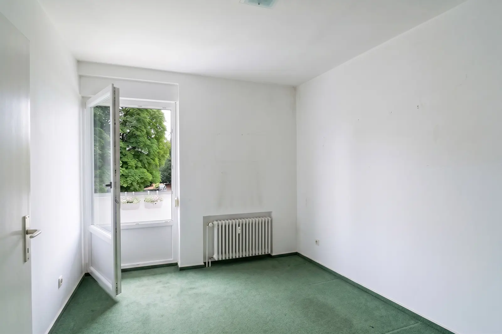 An empty white room with green carpet, a radiator and an open window through which trees and a balcony can be seen.