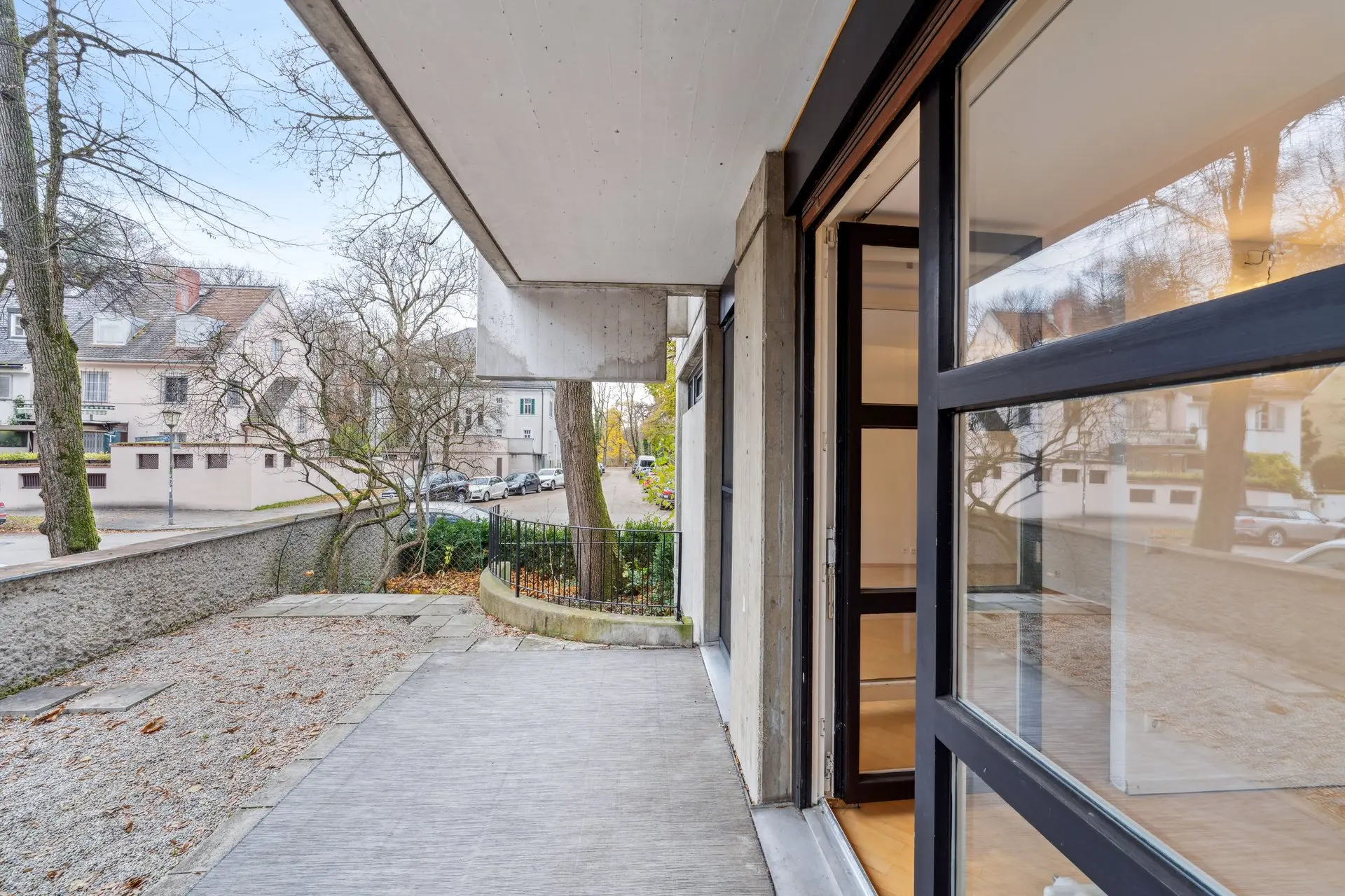 View from a modern building's balcony overlooking a residential street with trees and parked cars. Large glass door opens to the exterior.