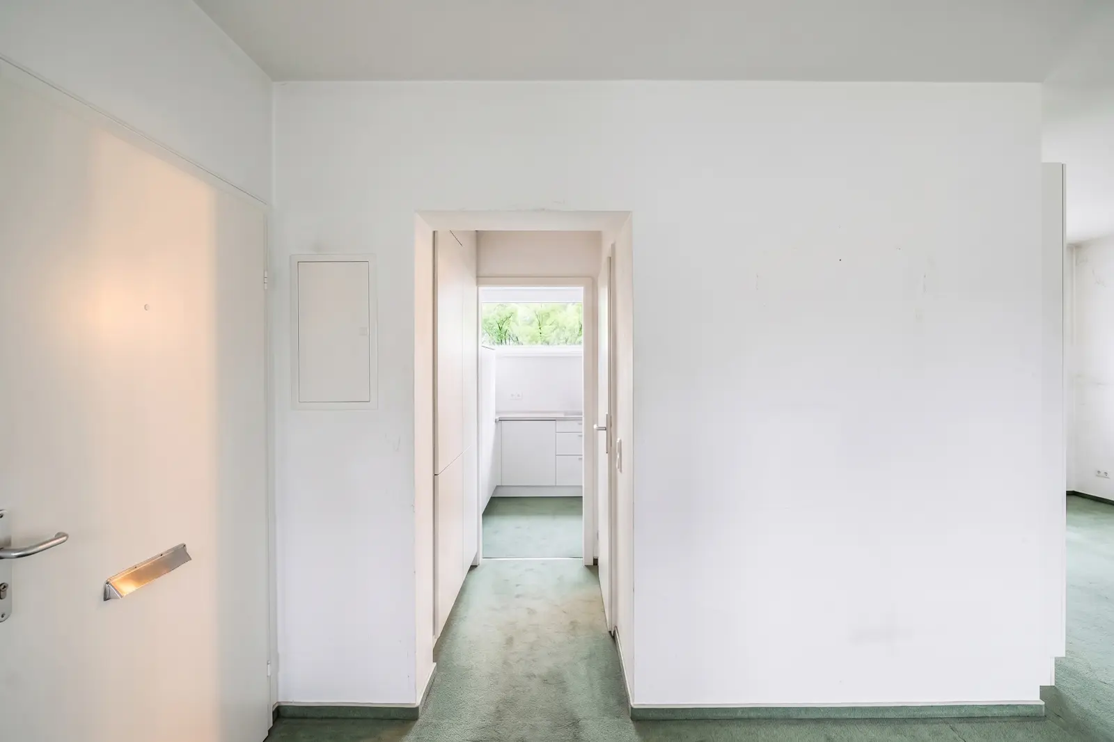 View of a white hallway with light green carpet leading to a small kitchen with a window at the far end. The hallway has plain walls and minimal decoration.