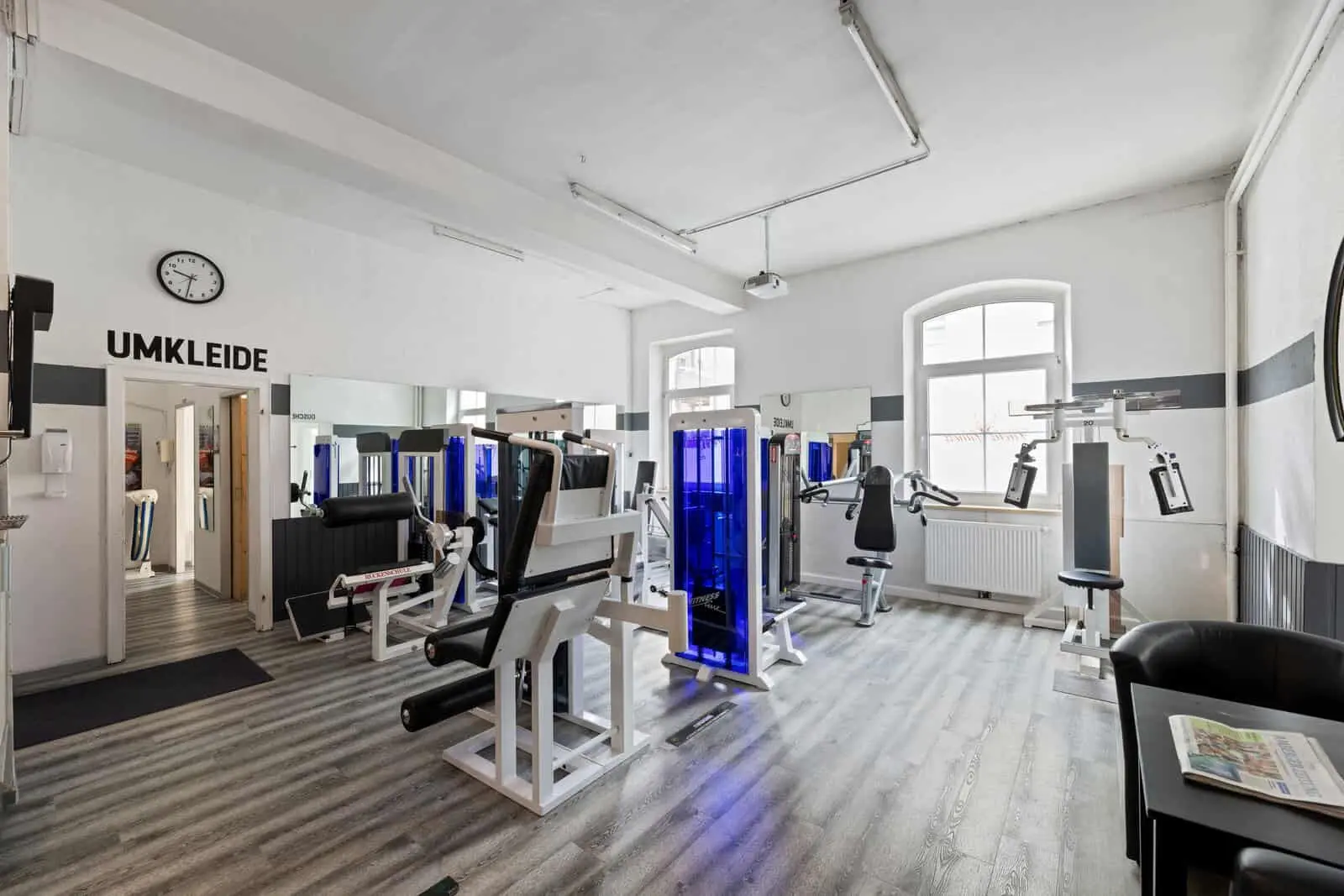 A gym with various weight machines, mirrors on the walls, a clock above a door labelled “Umkleide” (changing room) and a chair with newspapers on a side table.