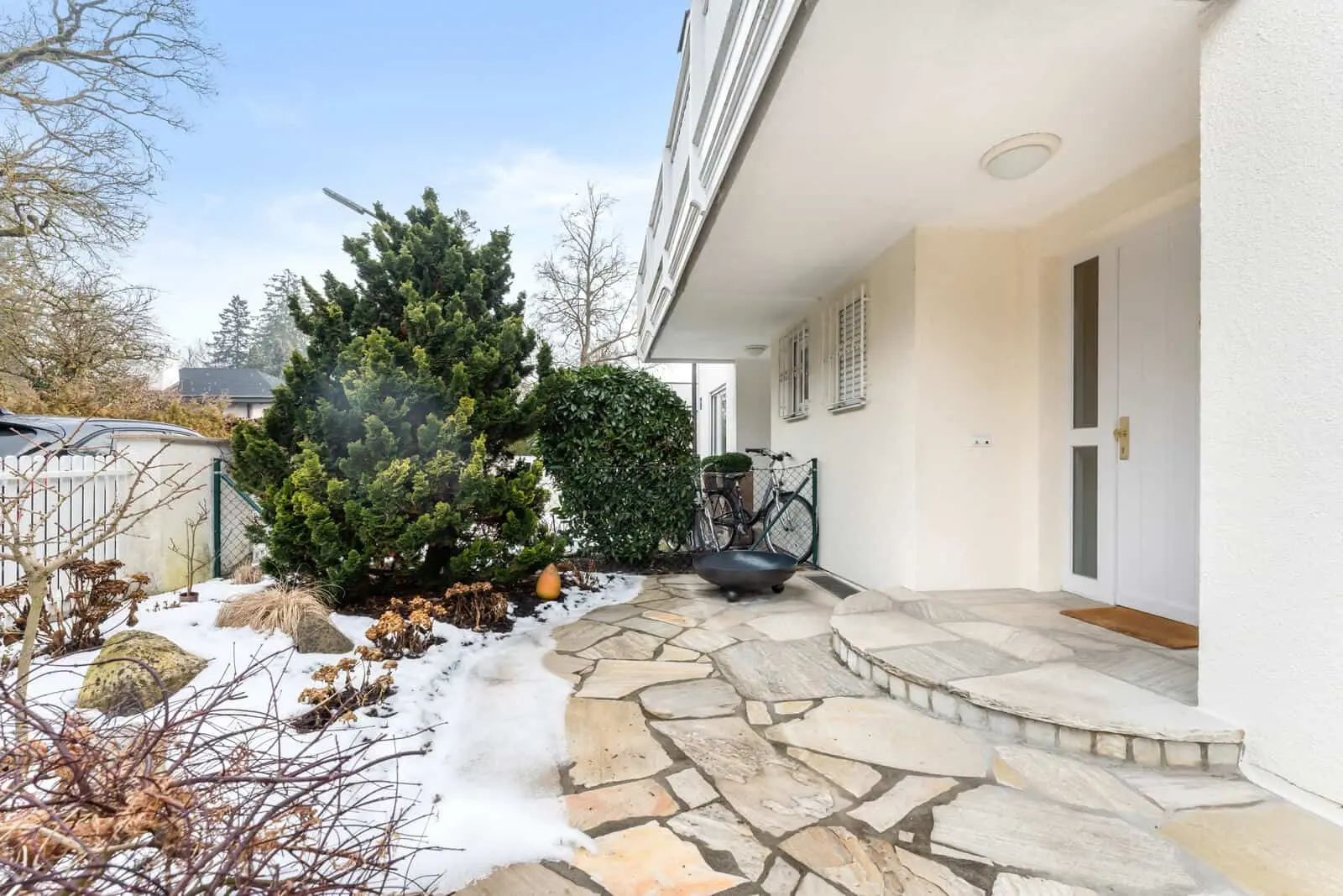 A stone walkway leads to a white house entrance, partially covered by a roof. Some snow remains on the ground, and bicycles are parked near the door, with bushes on either side.