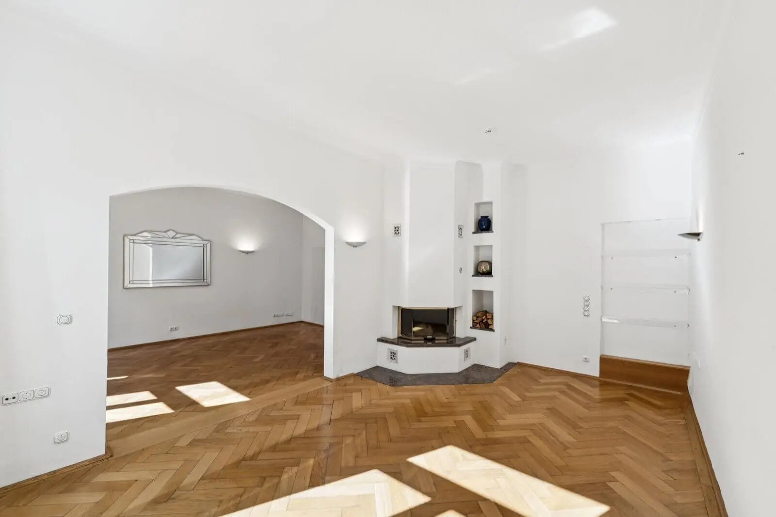 Empty room with white walls, parquet flooring, built-in corner fireplace, arched doorway, wall shelves, and a large mirror on the far wall.