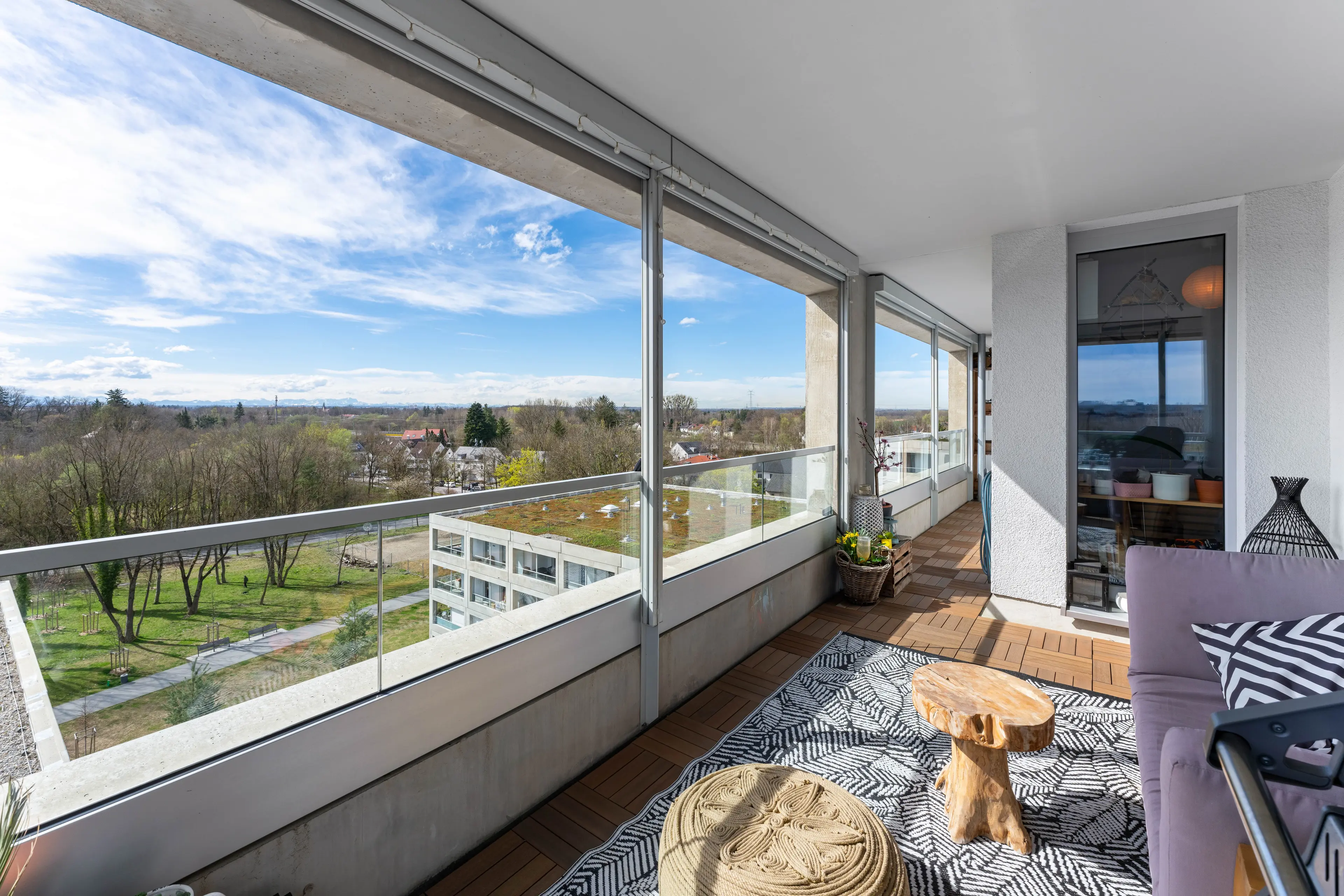 A modern balcony with large glass panels offers a panoramic view of a cityscape and park on a clear day. The space features a patterned rug, small table, pouf, and decorative items.