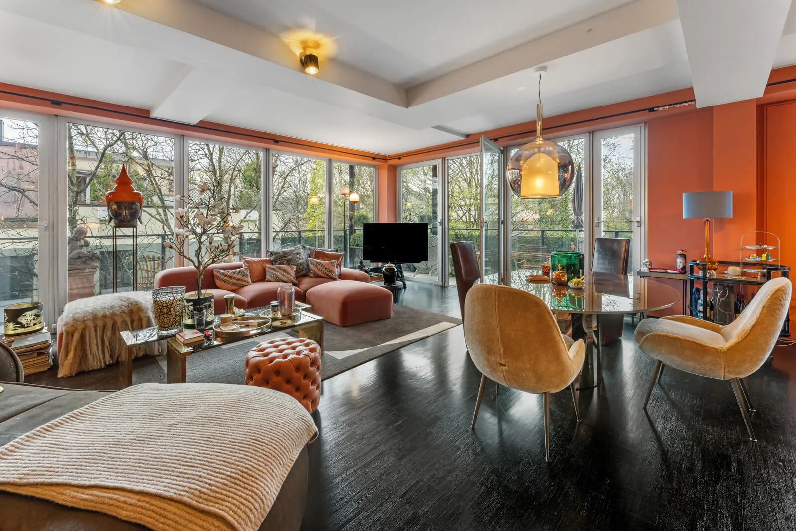A modern living room with large windows, orange walls, dark flooring, and a mix of seating including chairs and sofas. A television is central, with various decor items and a dining area in view.
