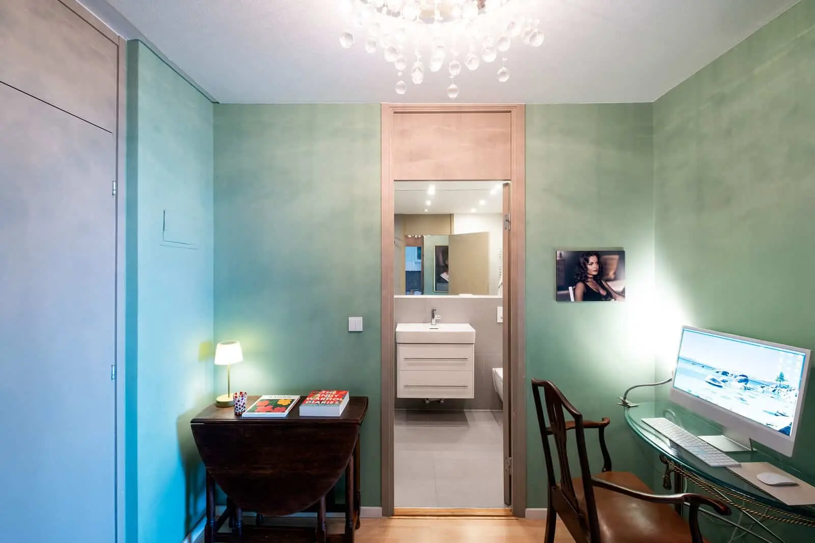 A small room with green walls, a wooden table with books and a lamp, a chair, and a desk with a computer; a door opens to a bathroom with a sink and mirror.