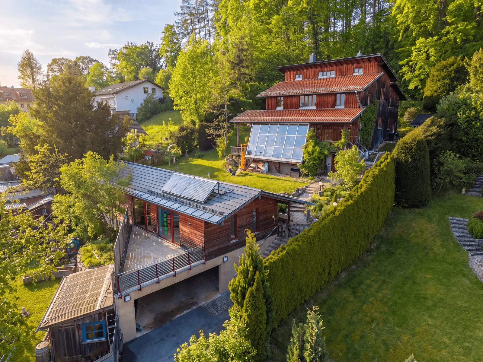 Two modern wooden houses with solar panels on the roofs are surrounded by trees and greenery on a hillside under a clear sky.