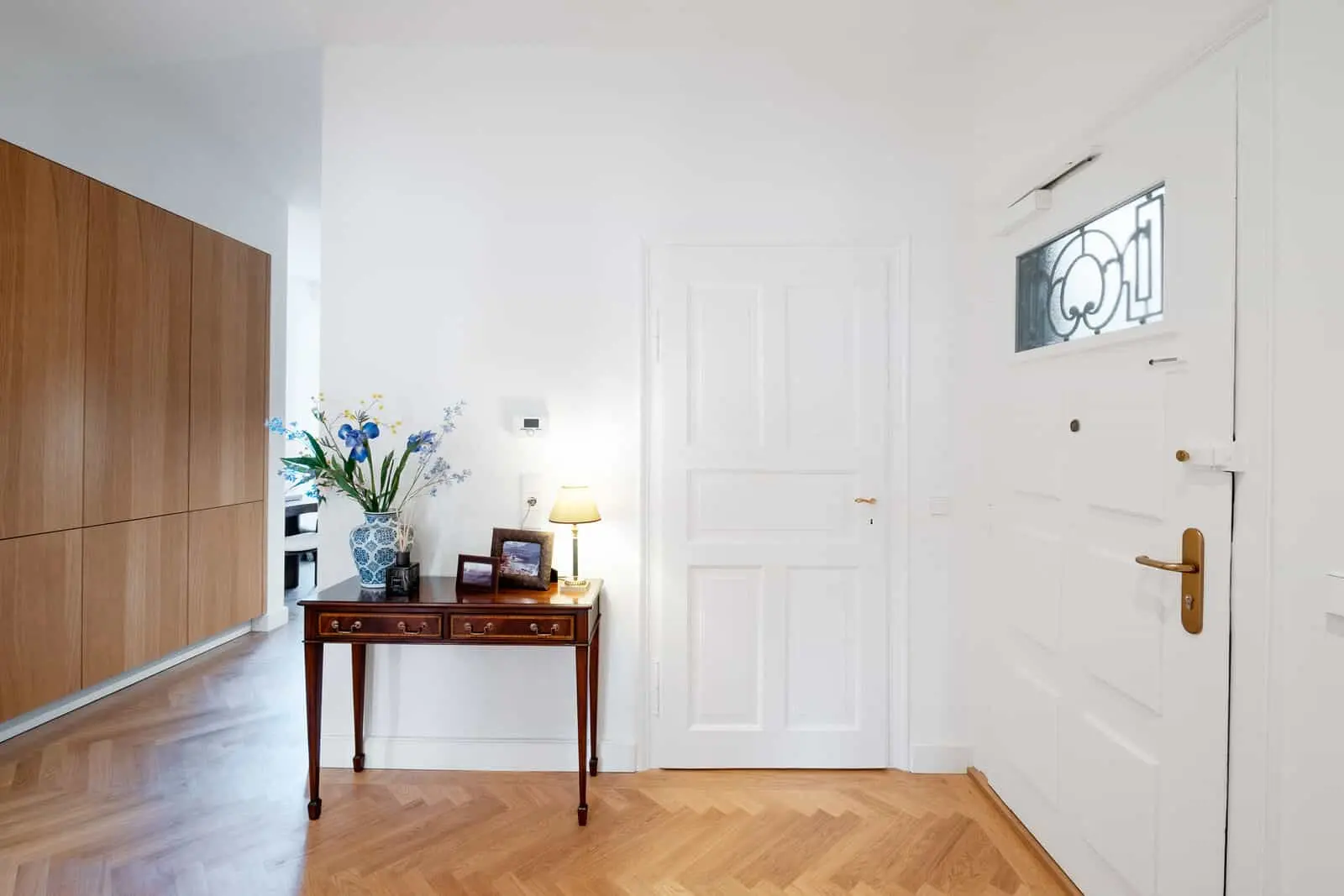 A bright entryway with a white door, double white interior doors, a wooden console table with flowers, photos, and a lamp, and built-in wooden cabinets on the left.