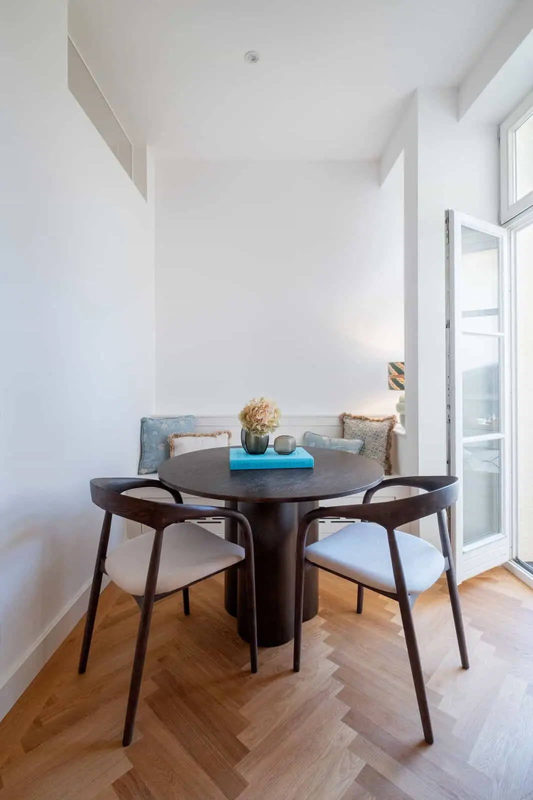 A small dining area with a round dark wood table, three chairs, light wood herringbone floor, large window, and pastel decor accents.