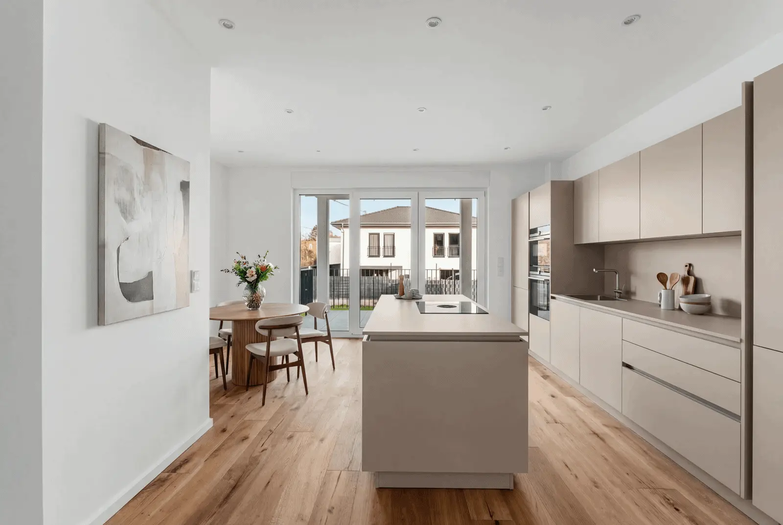 Modern kitchen with beige cabinets, a central island, built-in appliances, a wooden dining table with chairs, abstract wall art, and large windows overlooking a fenced backyard.