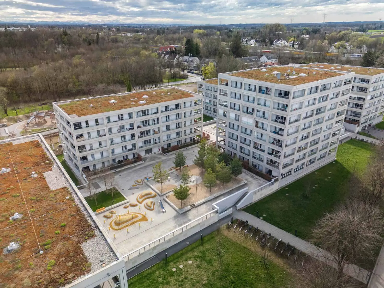Aerial view of modern apartment buildings with green roofs, a central courtyard with trees and benches, and surrounding grassy areas.