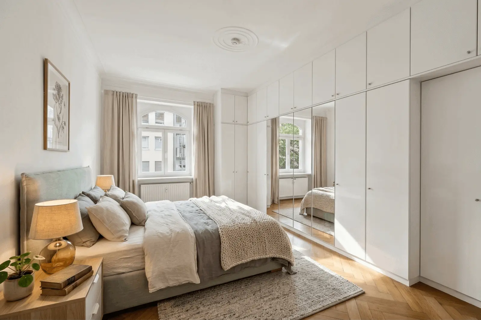 Bright bedroom with a neatly made double bed, bedside tables, large wardrobe with mirrored doors, wood floor, and two windows with curtains letting in natural light.