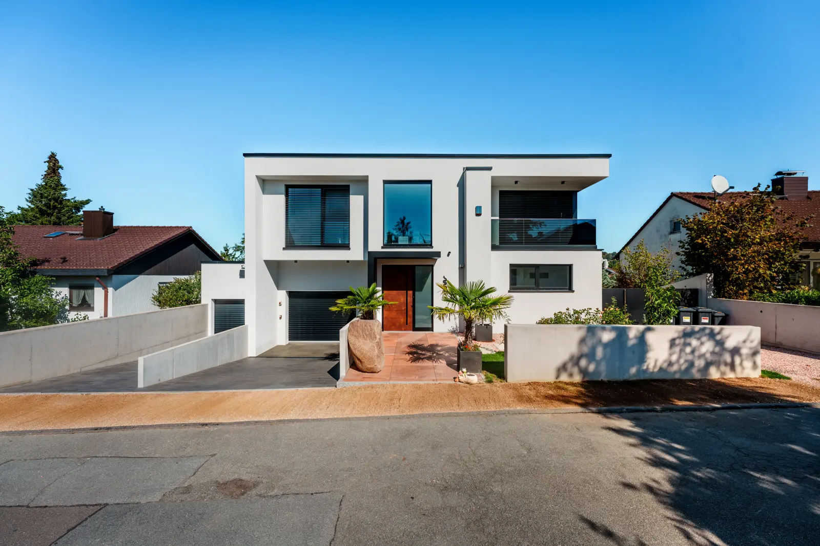 Modern two-story house with a flat roof, white exterior, integrated garage, and a small front courtyard with two palm trees.