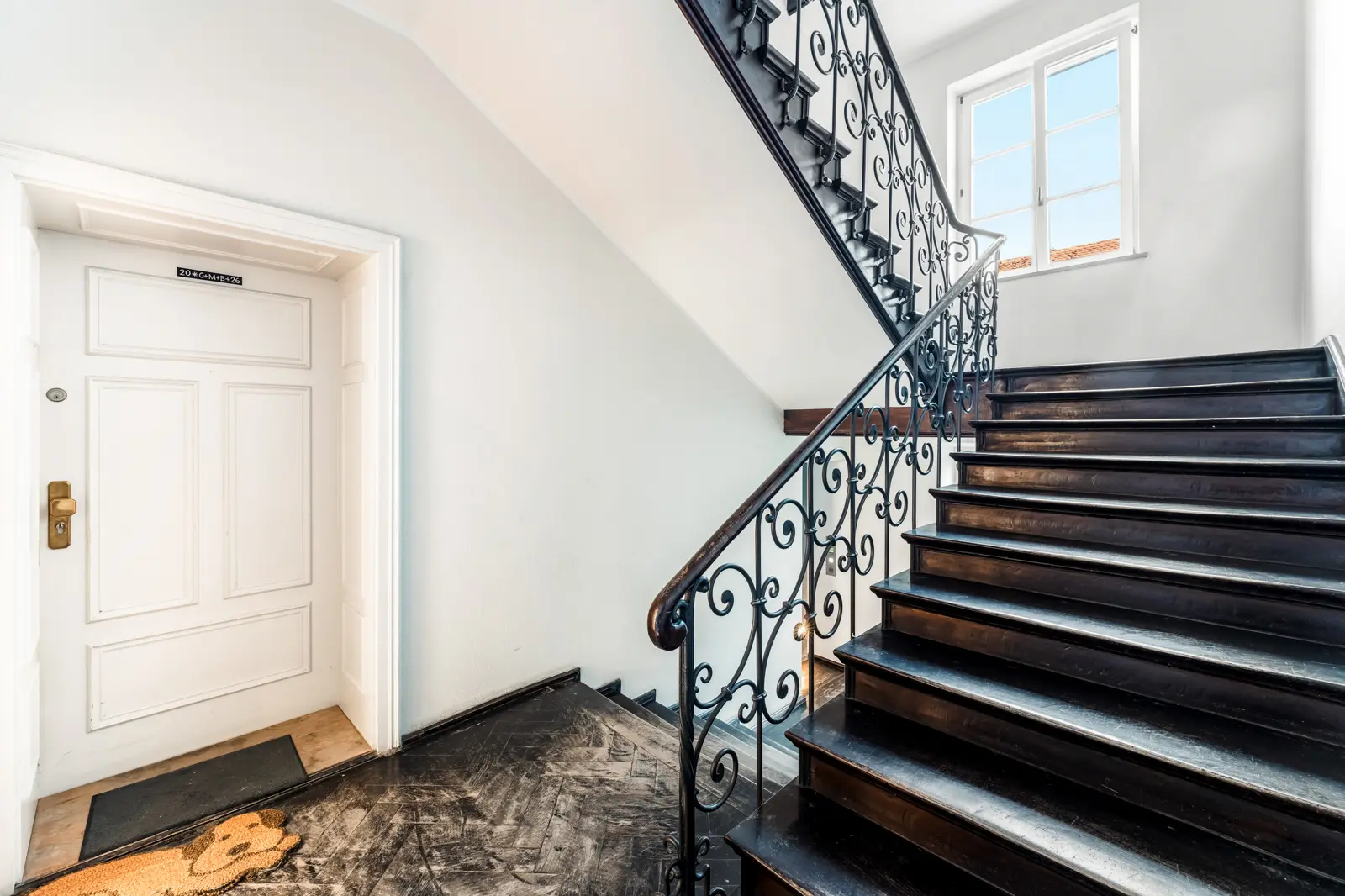 A black staircase with ornate railing next to a white door in a brightly lit hallway with a window at the landing.
