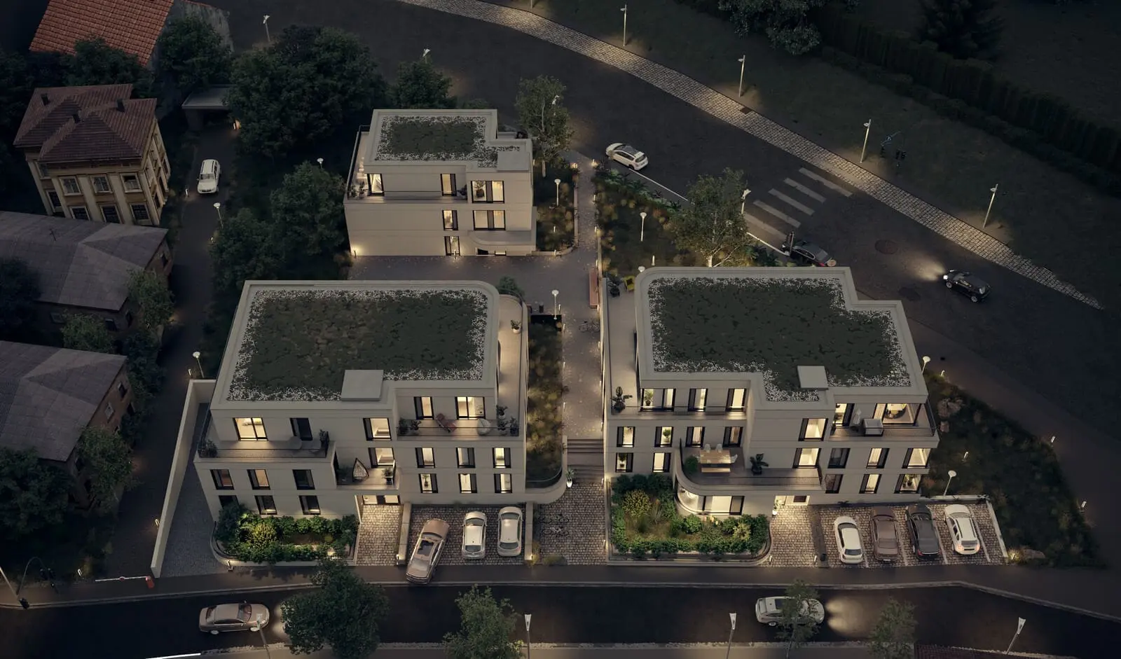 Aerial view of three modern apartment buildings at night, with illuminated windows, parked cars, and surrounding greenery.