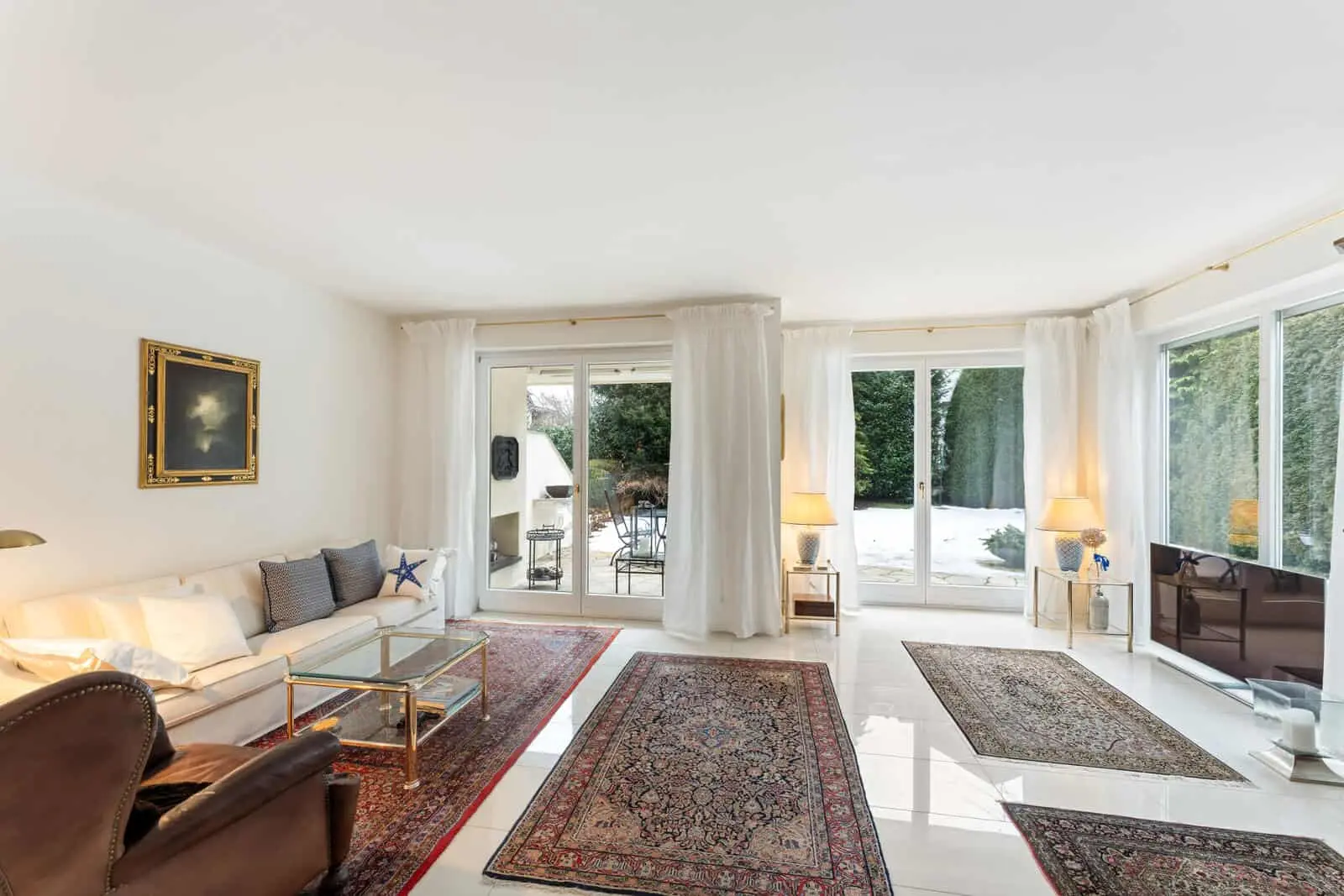 Bright living room with large windows, white walls, two area rugs, a sofa, glass coffee tables, armchair, lamps, and a view of a snowy outdoor patio.