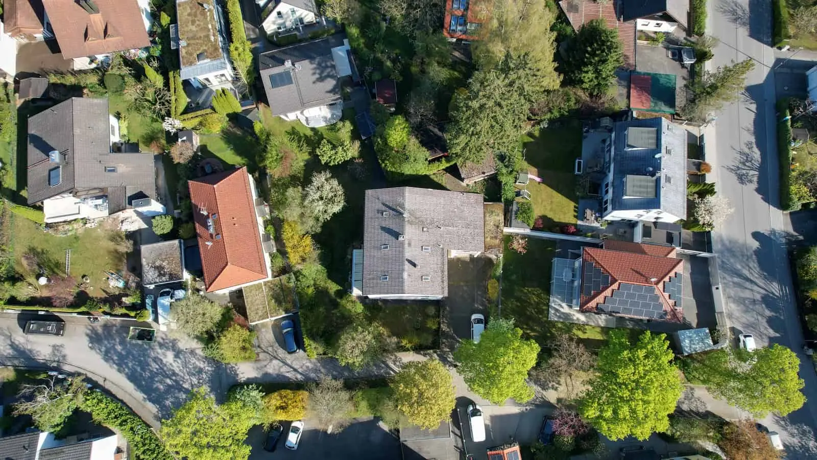 Aerial view of a residential neighborhood with houses, gardens, trees, parked cars, and streets on a clear day.