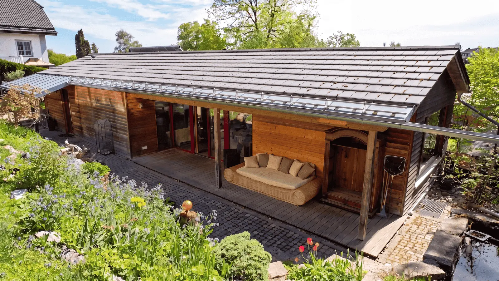 Single-storey wooden house with a covered veranda, outdoor seating and a garden with a variety of flowers and plants in front.