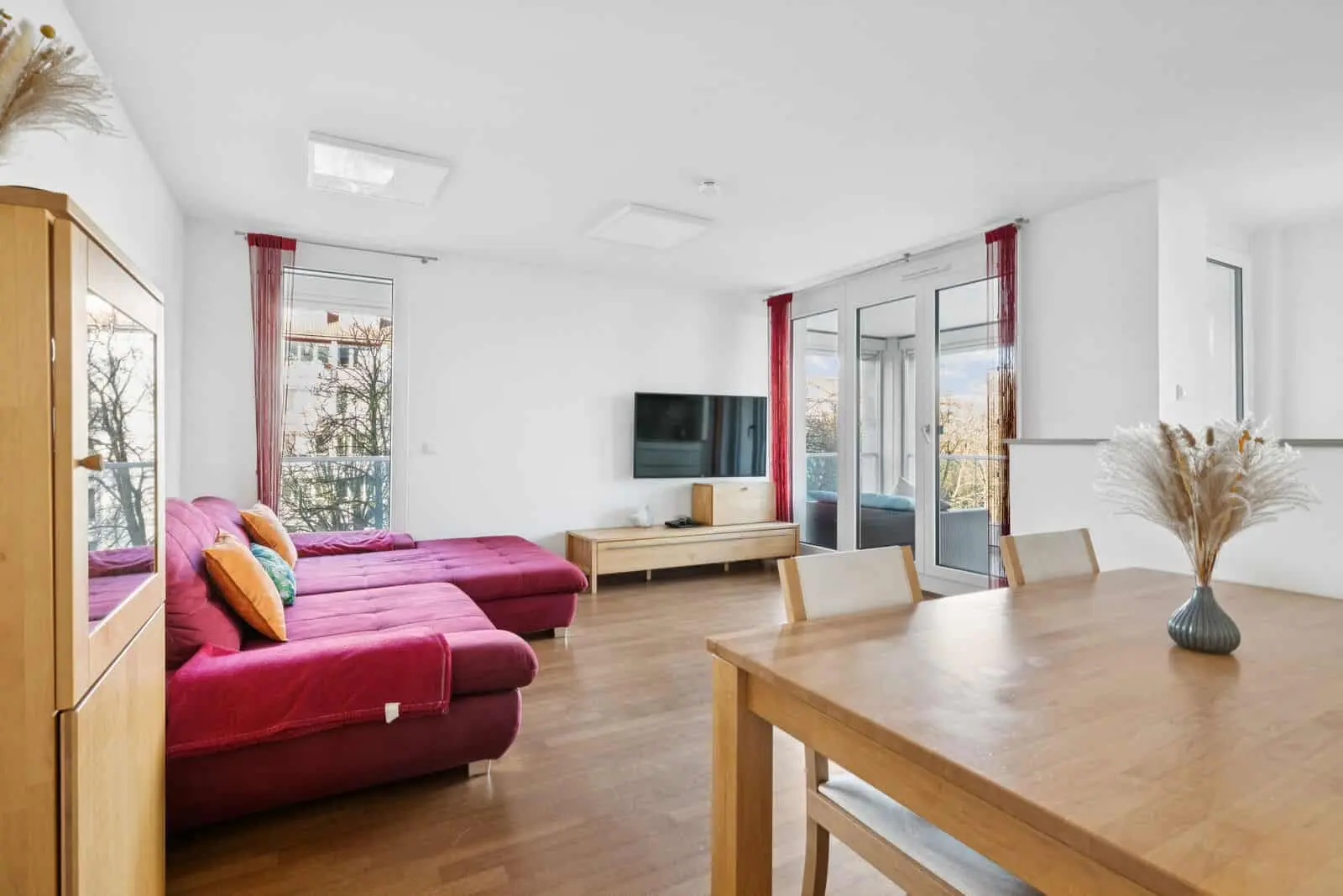Modern living and dining area with wooden furniture, a red sectional sofa, wall-mounted TV, large windows, and glass doors opening to a balcony.