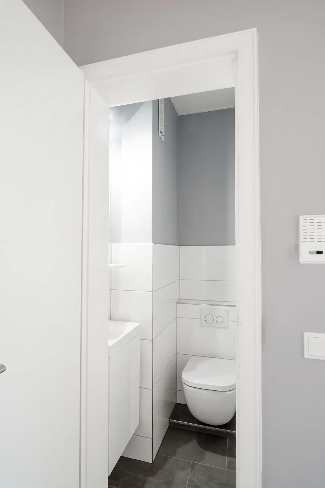 A small, modern bathroom with a wall-hung toilet, white tiles and grey walls, seen through a partly open door.