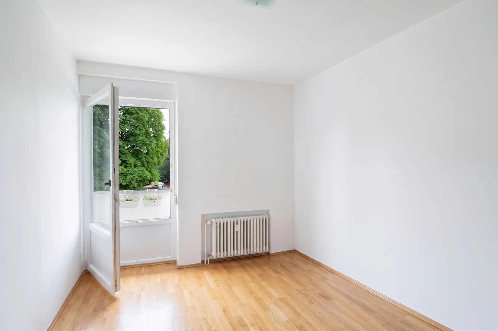 An empty white room with a wooden floor, an open window, a radiator and a view of green trees outside.