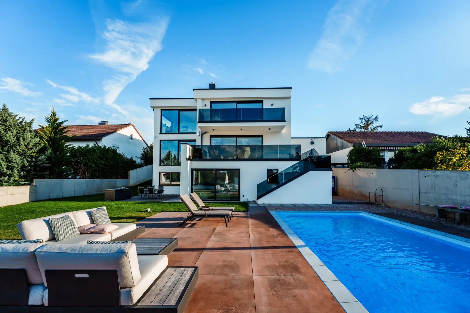Modern three-story white house with balconies, a patio with lounge furniture, and a rectangular swimming pool.