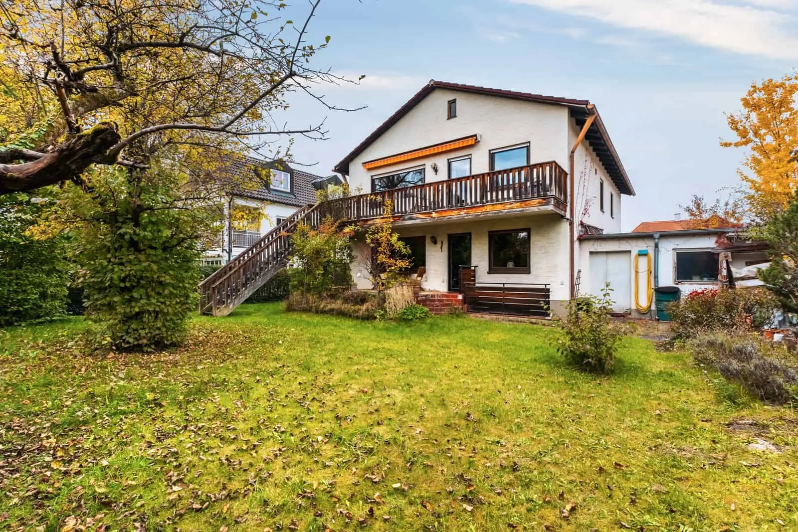 Two-story white house with a wooden balcony and exterior staircase, surrounded by a grassy yard with trees and autumn leaves.