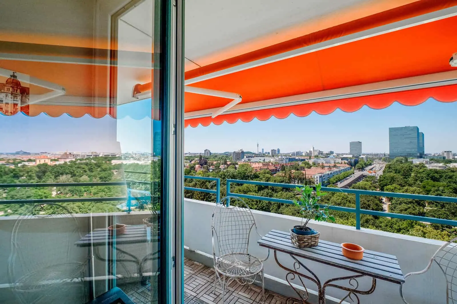View from a balcony with metal chairs, a small table, potted plants, and an orange awning, overlooking a cityscape and green park on a clear day.