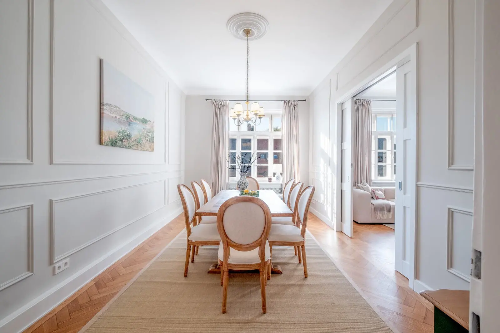 Bright dining room with a wooden table, six chairs, chandelier, landscape painting on the wall, large window, and view into an adjacent living room.