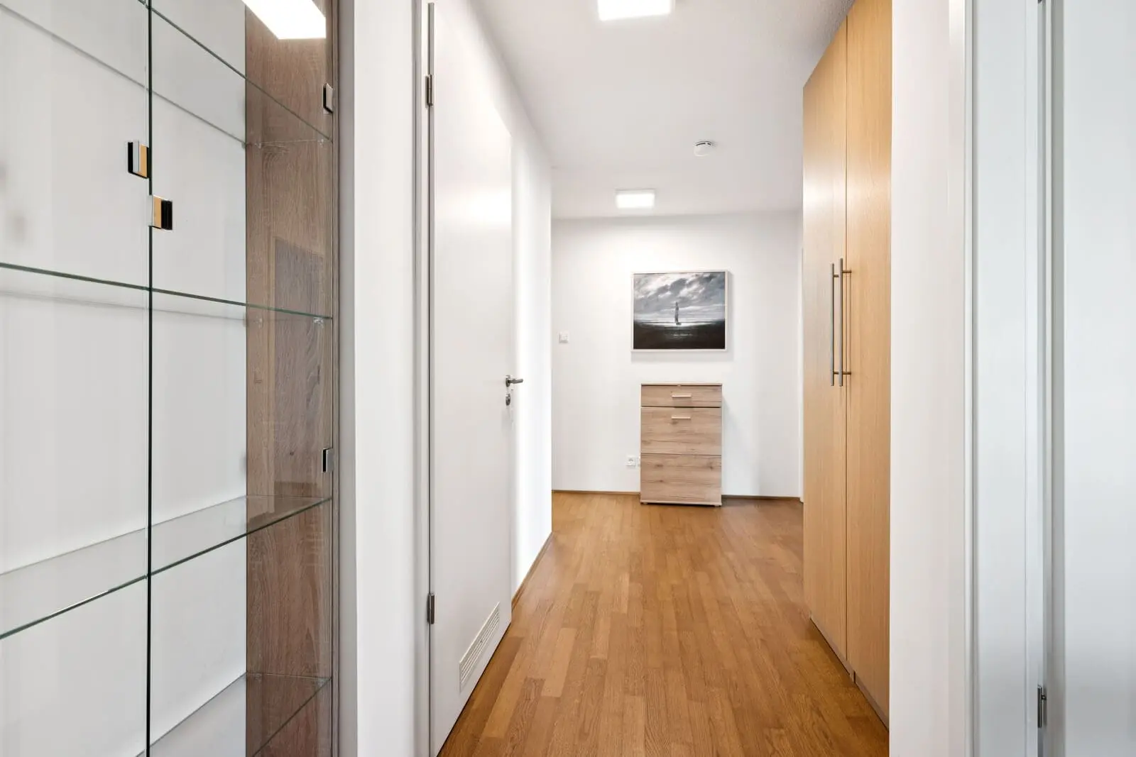 A hallway with wood flooring, a glass display cabinet on the left, white doors, built-in wooden closets on the right, and a small dresser with a framed photo at the end.
