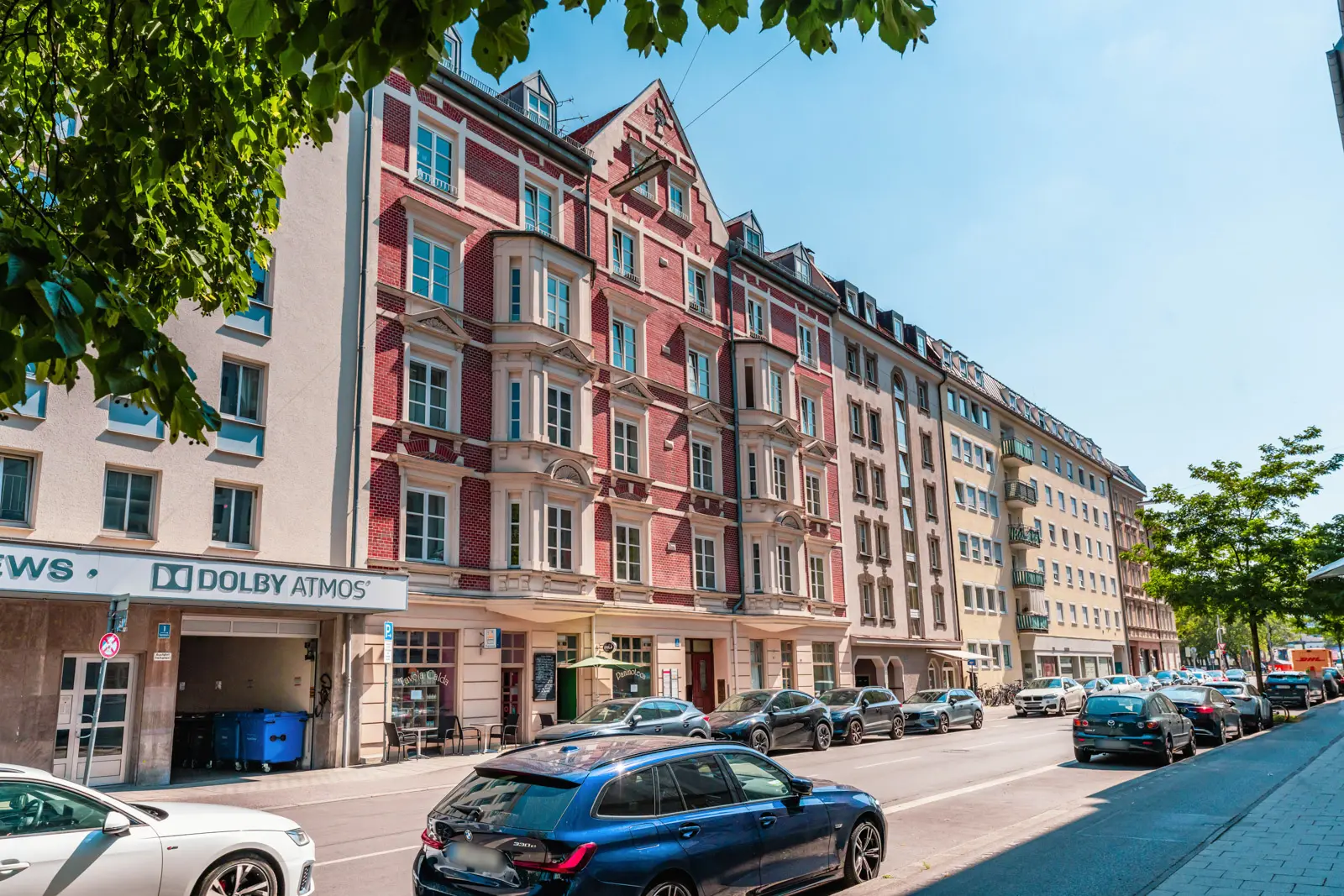 Street view of parked cars in front of multi-story residential and commercial buildings on a sunny day, with trees partially framing the scene.