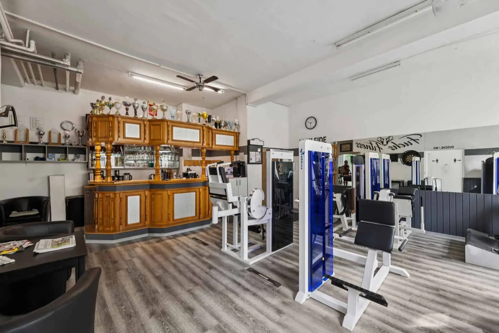 A gym with fitness equipment in the foreground and a wooden counter with trophies and mirrors in the background.