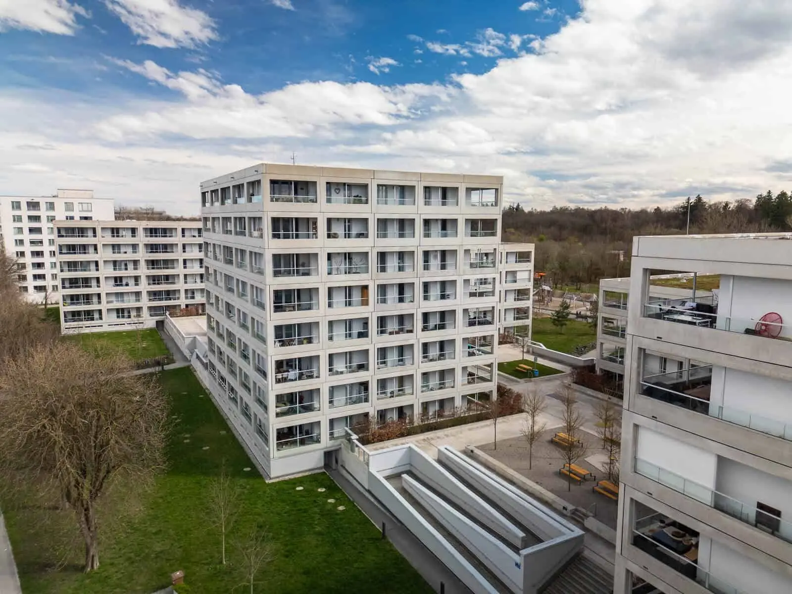 Modern apartment buildings with multiple floors, flat roofs, and balconies, surrounded by green lawns and trees under a partly cloudy sky.