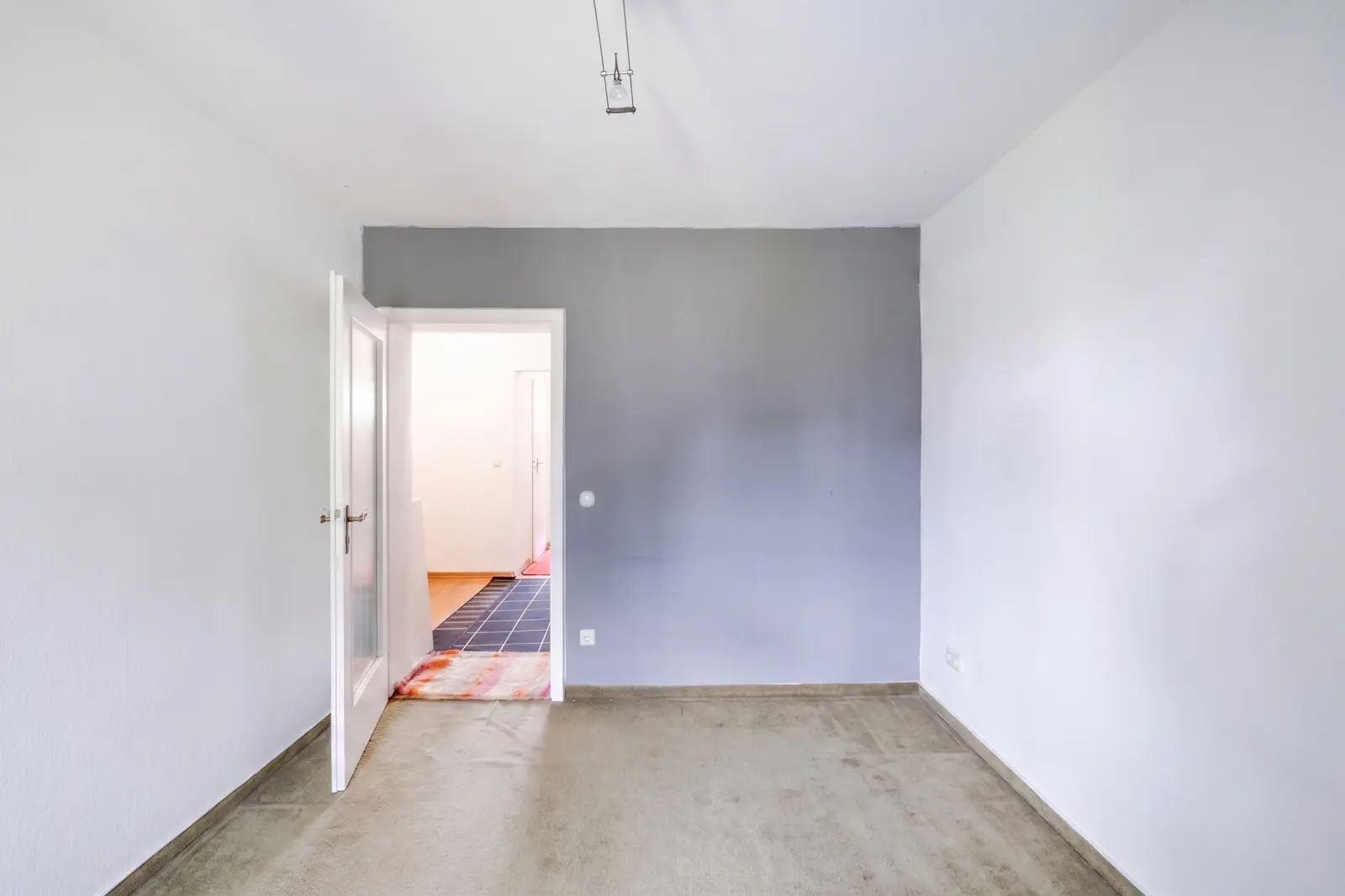 An empty room with grey carpet, a grey accent wall, an open white door and a view of a brightly lit hallway with a tiled floor and another door.