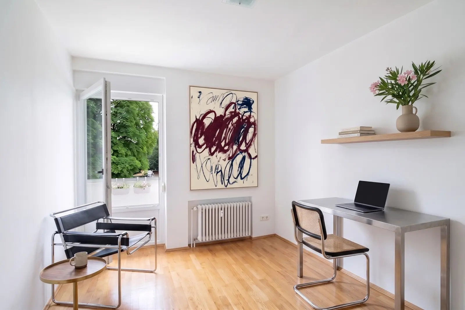 Minimalist home office with a desk, a laptop, a chair, two armchairs, a coffee cup, abstract wall art, a floating shelf with books and flowers, and an open door leading to a balcony.