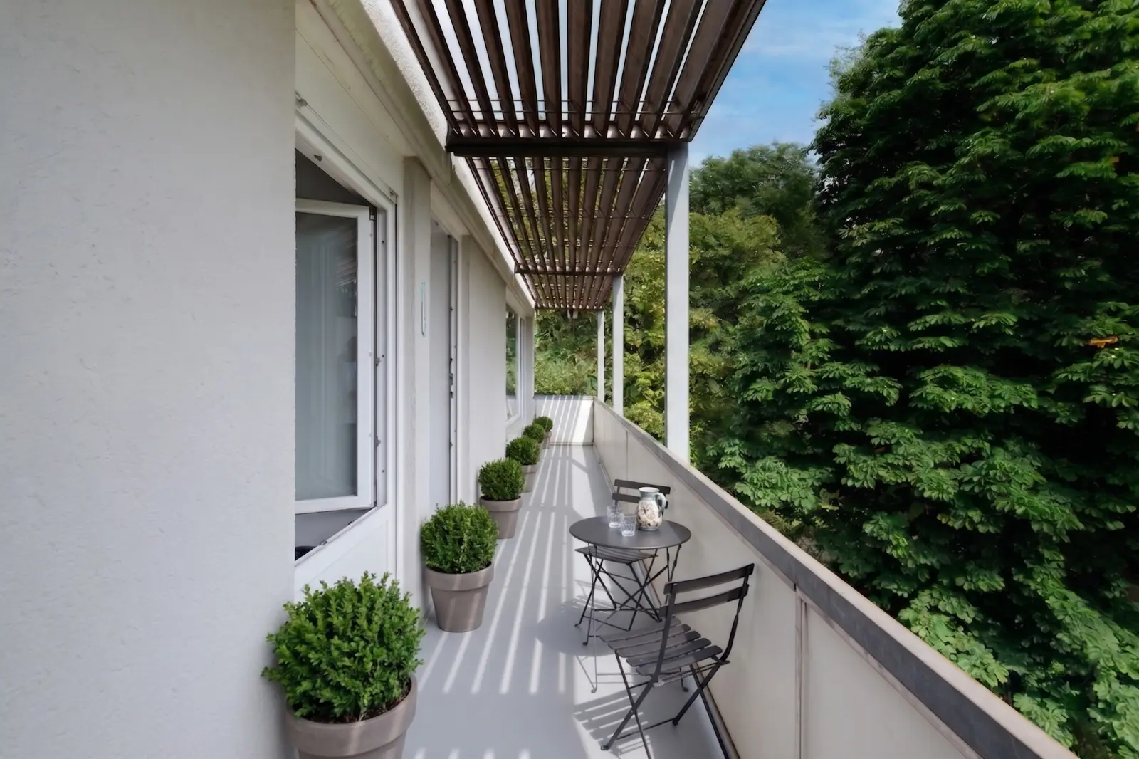 A long balcony with potted plants, a small round table, two chairs and a coffee cup, overlooking lush green trees under a louvred roof.
