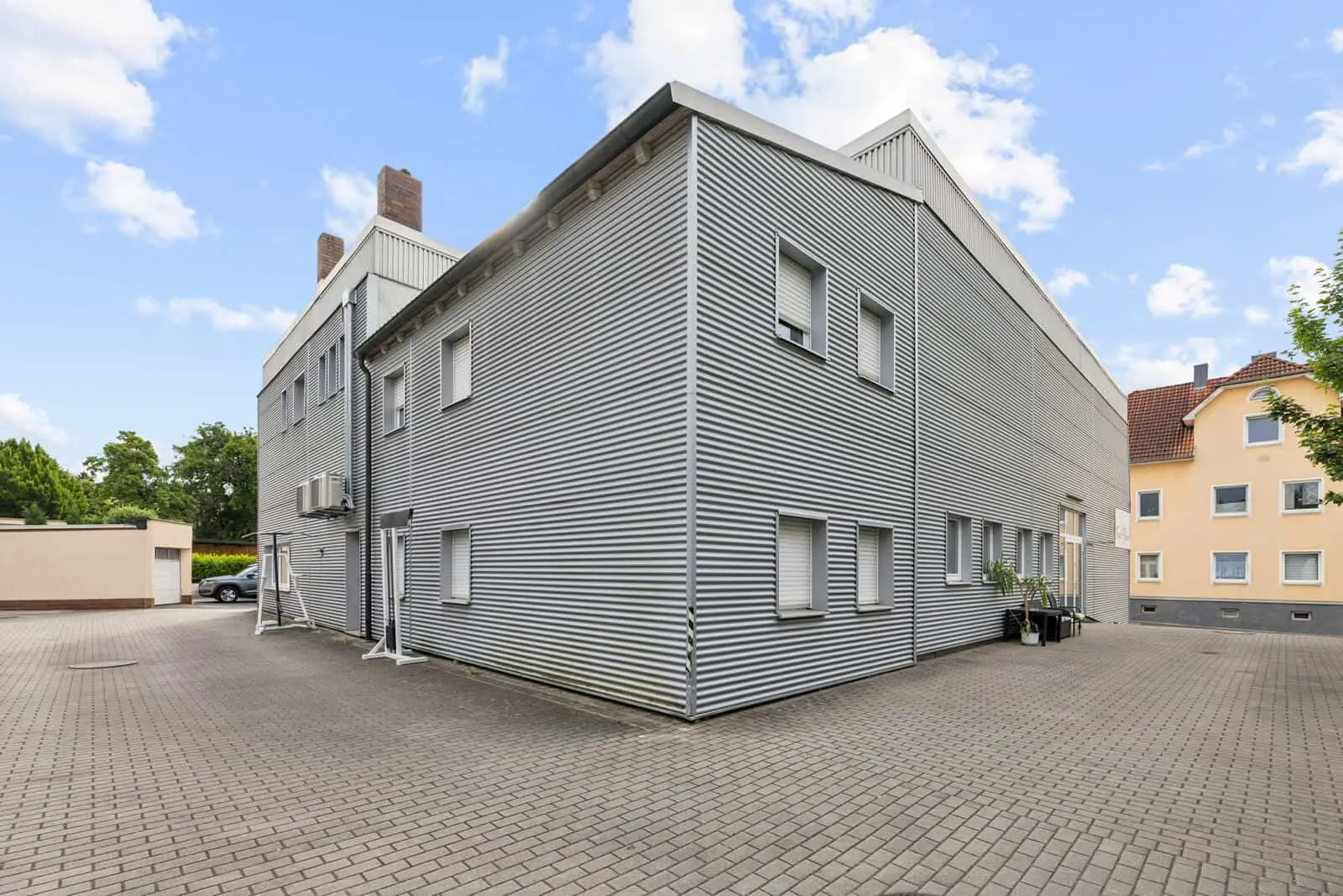 A modern industrial building with corrugated metal cladding, a flat roof and several closed windows, surrounded by a paved car park under a partly cloudy sky.