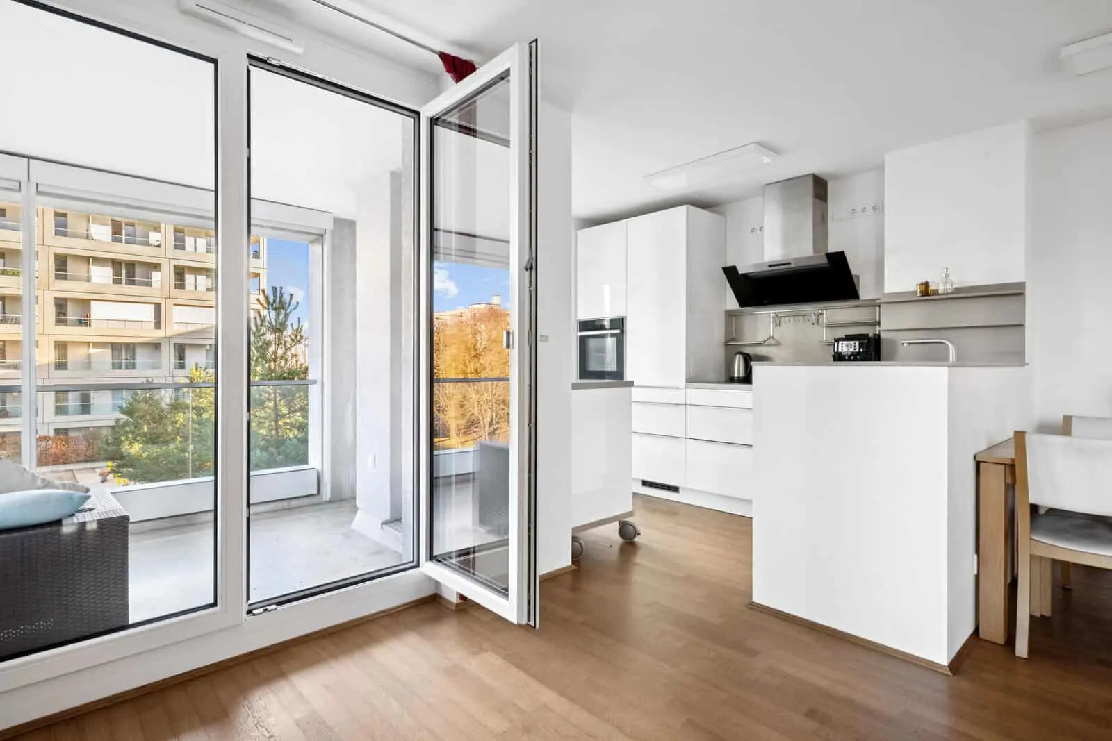 Modern white kitchen with wooden floor, open glass door leading to a balcony with outdoor seating, and view of apartment buildings and trees outside.