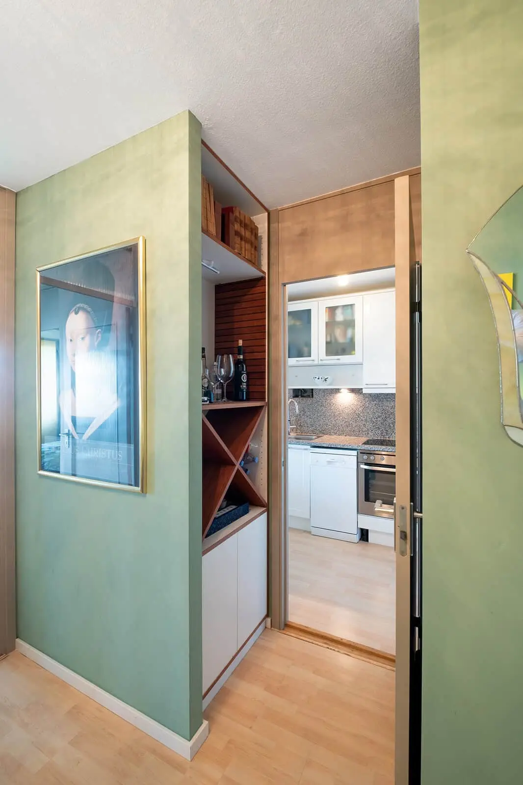 A small kitchen with white cabinets is seen through a doorway; the foreground features green walls, a wooden floor, a wall shelf with wine storage, and a framed portrait.