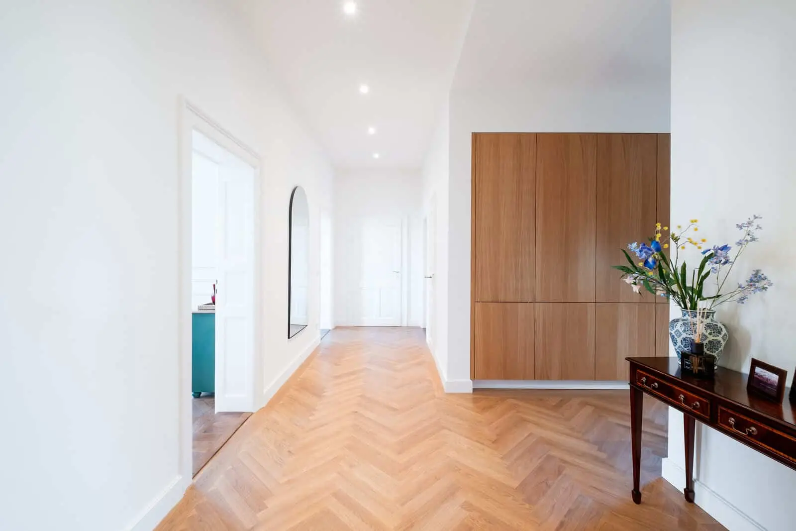 Bright hallway with herringbone wood flooring, white walls, a large mirror, wooden cabinets, and a table with a vase of flowers and framed photo.