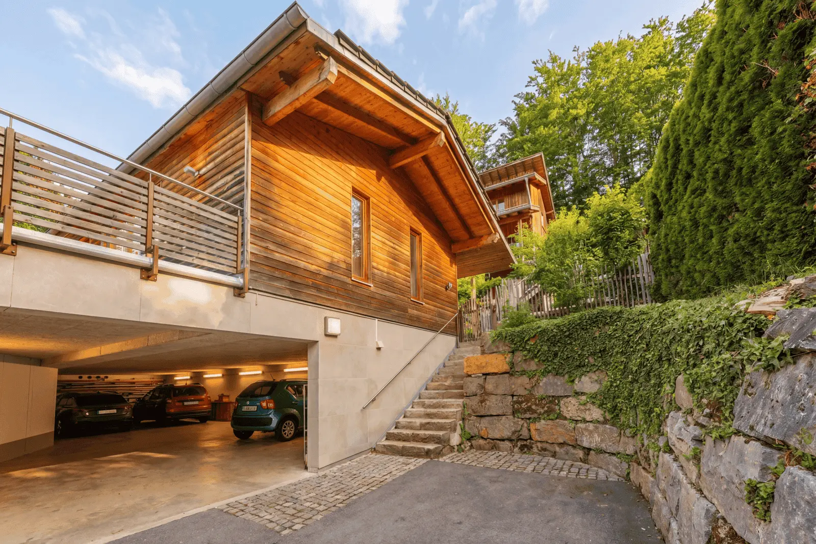A modern wooden house with an open garage below, parked cars, a stone staircase leading up and green spaces around the property.