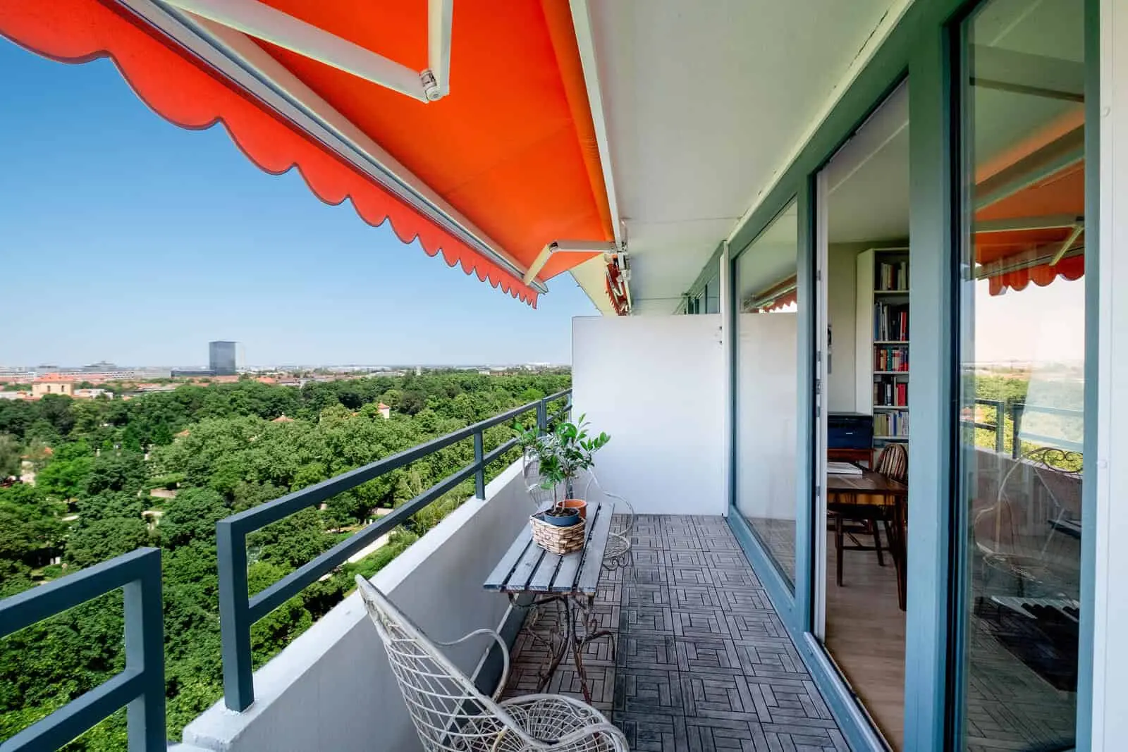 A modern apartment balcony with orange awnings, a small table and chair, potted plants, and a view of trees and buildings in the distance.