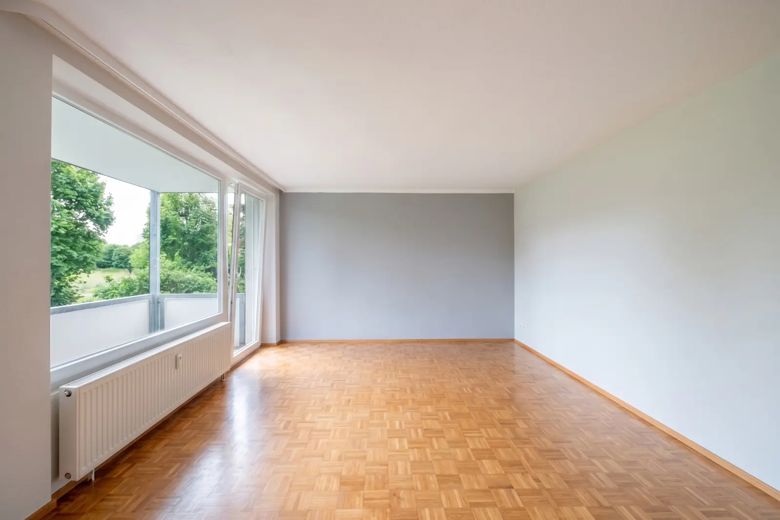 An empty living room with parquet flooring, a large window, white walls, a grey accent wall and a view of greenery.