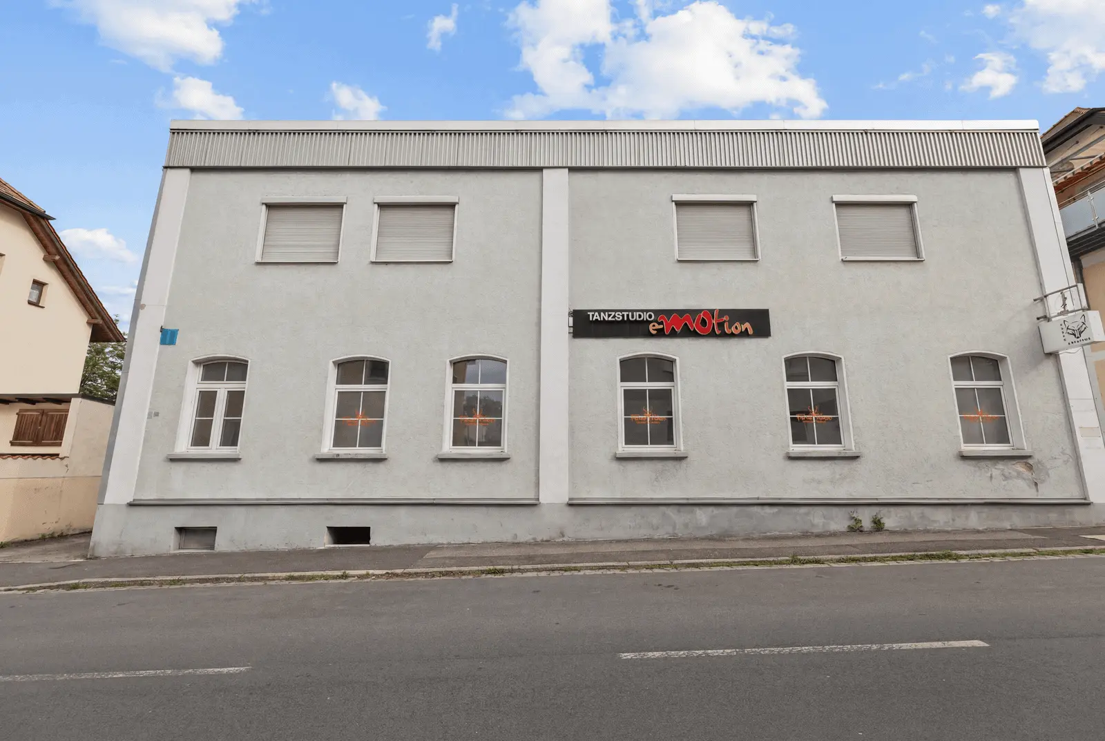 A light grey two-storey building with six closed windows on the upper floor, five windows on the lower floor and a sign reading “Tanzstudio eMotion” above the door.
