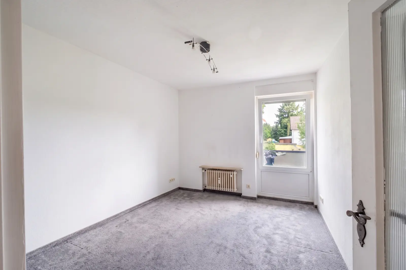 An empty room with white walls, grey carpet, a window letting in daylight, a radiator under the window and a partly open door on the right. There is a ceiling light.
