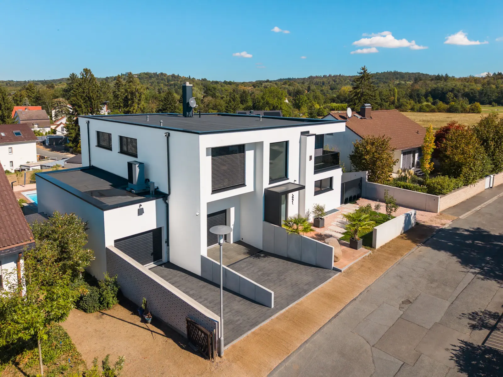 Aerial view of a two-story modern white house with a flat roof, dark window accents, and a paved driveway, surrounded by greenery.