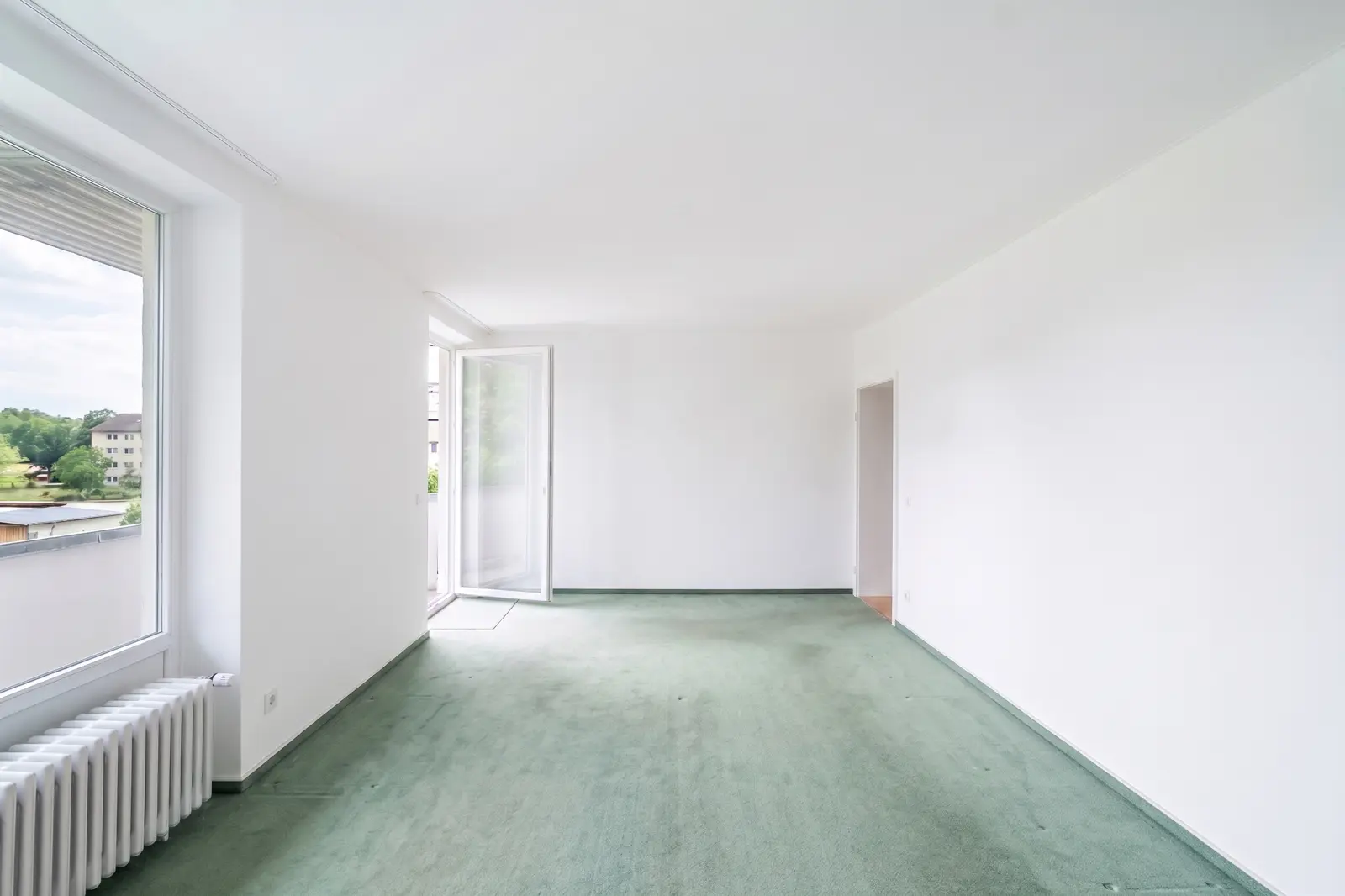An empty room with white walls, green carpet, a radiator under a window, an open door to the balcony and a door to another room.