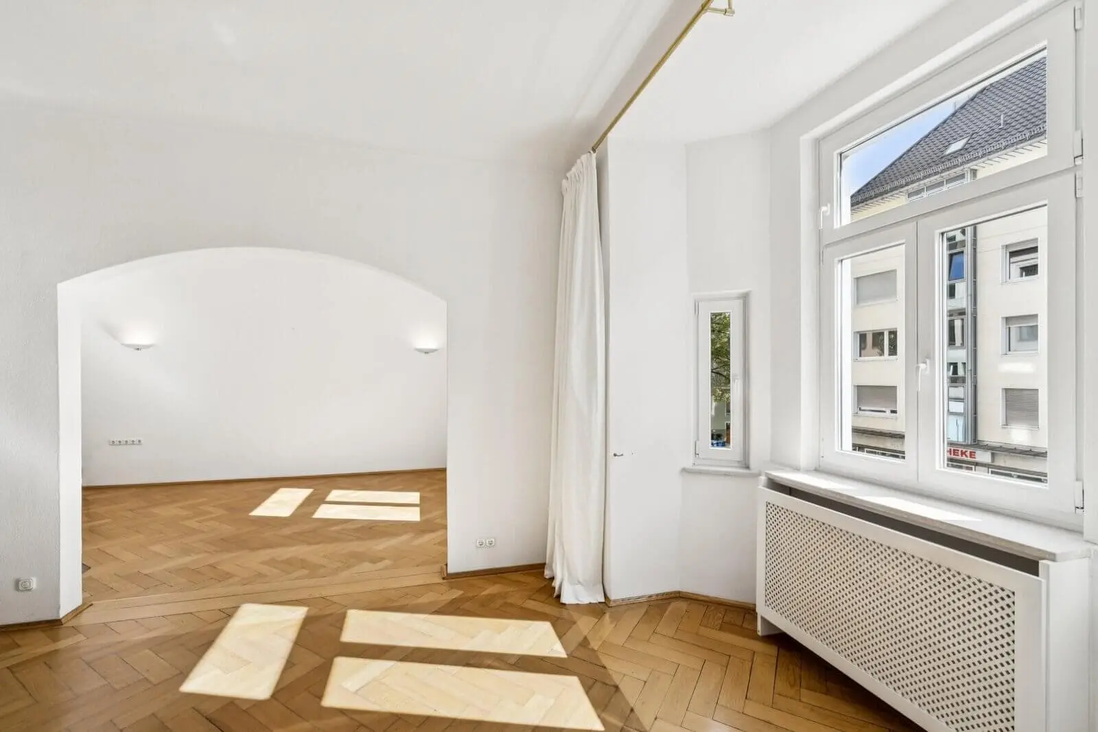 Bright, empty room with parquet flooring, large windows, white walls, and an open archway leading to another space. Sunlight casts shadows on the floor.