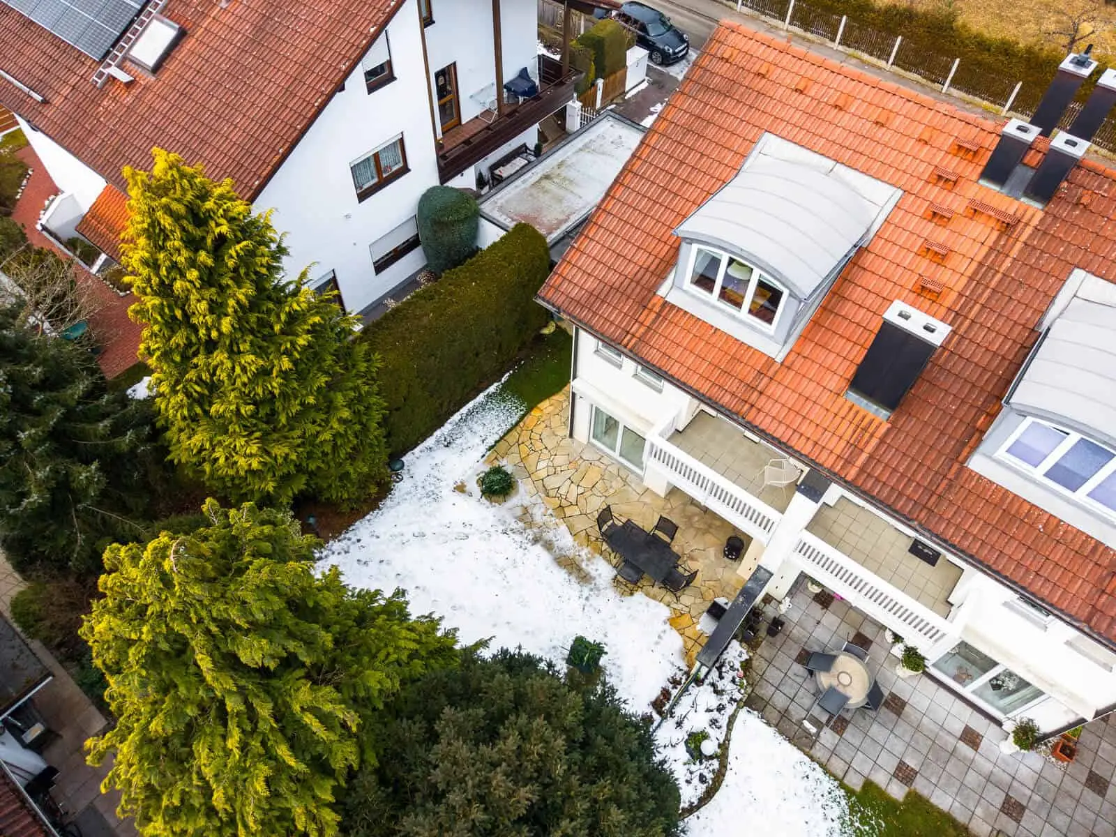 Aerial view of two houses with red-tiled roofs, a snow-dusted yard, trees, and a stone patio with outdoor furniture.